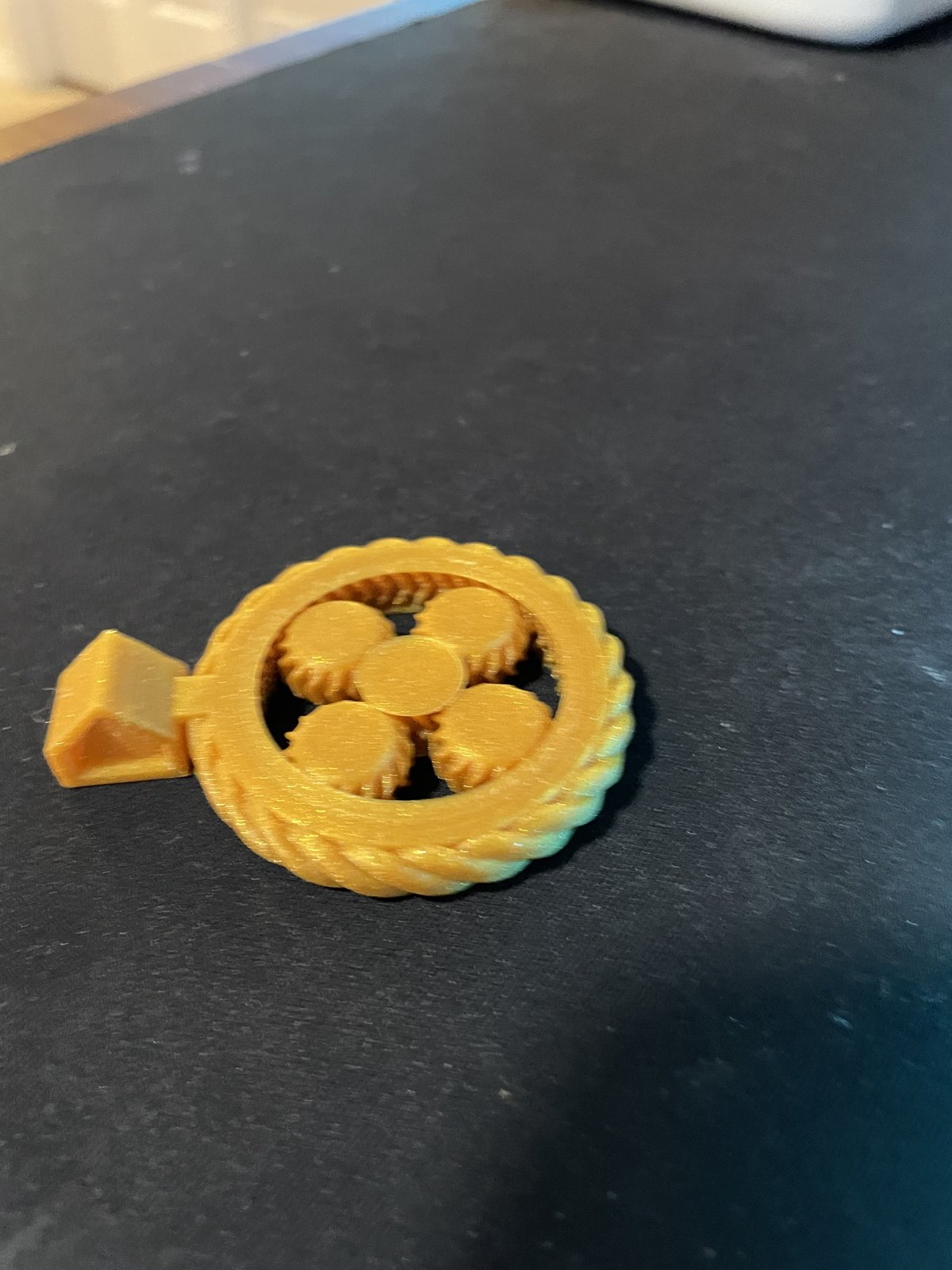 The printed gold v2 pendant on a dark mat: four planet gears around the sun inside a braided rim, with the chain bail
